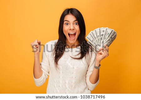 Portrait of an excited young woman holding money banknotes and celebrating isolated over yellow background