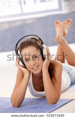 Attractive young female girl listening to music, using headphones, laying on floor at home, smiling.