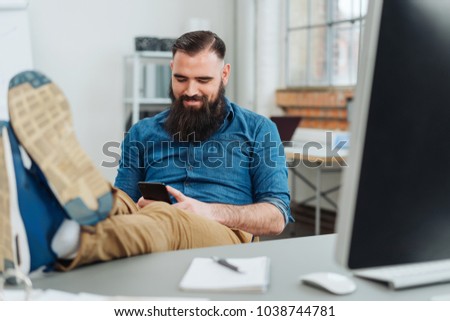 Relaxed casual businessman with his feet on the desk sitting back in his chair reading a text message on his mobile phone with a smile