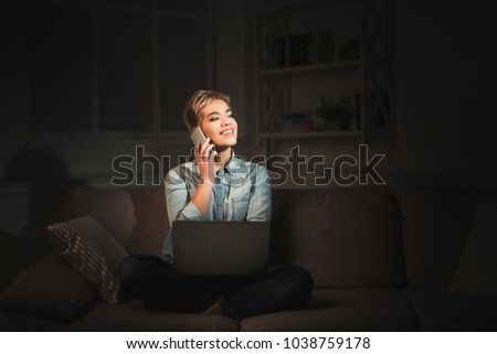 Happy young woman working late at home office, using laptop and talking on mobile, copy space. Casual girl in screen light in dark room background. Technology and overworking concept