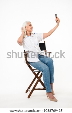 Full length portrait of a smiling mature woman sitting on a chair and taking a selfie isolated over white background