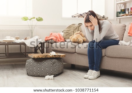 Desperate helpless woman sitting on sofa in messy living room. Young girl surrounded by many stack of clothes. Disorder and mess at home, copy space Royalty-Free Stock Photo #1038771154