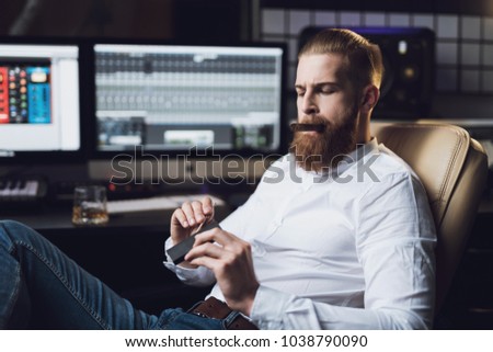 The male producer sits in the recording studio behind the remote control and smokes a cigar. He's wearing a posh white shirt. He has a very confident look.
