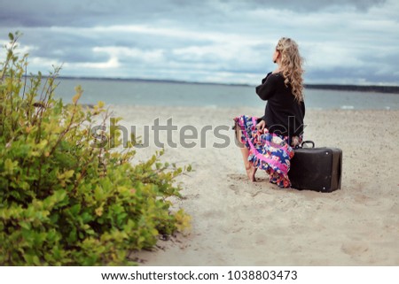 Young woman sitting on lonely beach and looking at the sea. Romantic picture. Beautiful girl waiting on a suitcase looking into the distance and sad