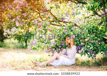 Young cute pretty blonde amazing model girl in white dress portrait in park outdoor. Beautiful happy girl with expressive face in blooming lilac garden. Spring is coming. Branches and trees. Easiness.
