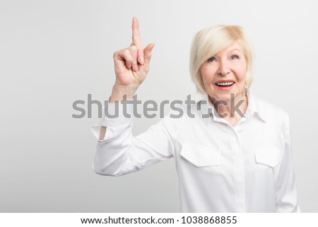 Nice picture of old woman standing at the white wall and pointing up. A brilliant idea has come up to her mind. Now she wants to materialize it. Isolated on white background