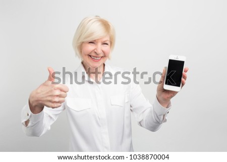 A picture of mature woman with new smartphone. She has tested it and admitted this phone is a good one. That's why she shows a big thumb up. Isolated on white background.