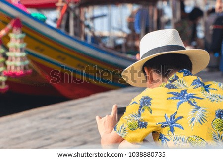 View from back of young man wearing vintage style shirt using smartphone to take a photo with motorboat at harbor. Travel in Chantaburi Province, Thailand. Soft focus and blur. Tourist concept.