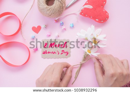 Woman hand holding white flowers with writing Happy International Women's Day message on white paper with handmade red heart, beautiful flowers paper stars and a lot of wooden heart on pink background