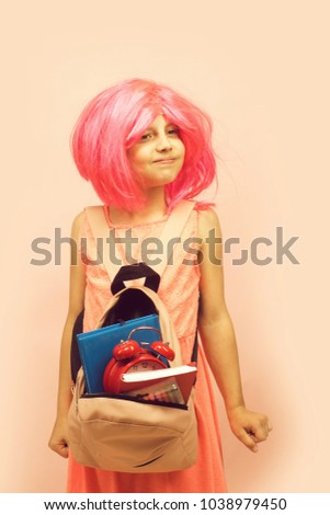 Girl with wig in pink dress with backpack. Back to school and learning concept. Pupil with funny face expression, isolated on light pink background. Kid with alarm clock and books.