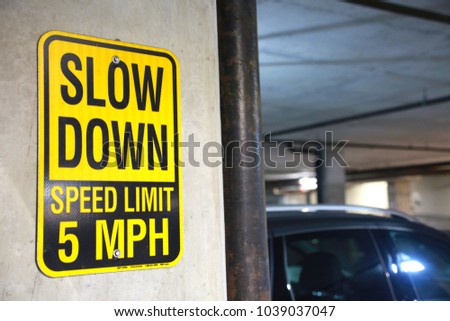 Yellow and Black Slow Down Speed Limit 5 Mph Sign in a Parking Garage with Cars Out of Focus in the Background