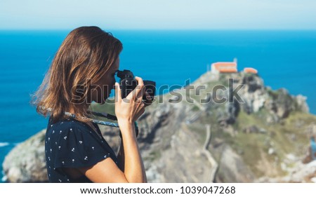 Tourist traveler photographer making pictures sea scape on photo camera on background ocean gaztelugatxe steps, hipster girl looking on nature horizon, relax holiday, blank space blue waves view