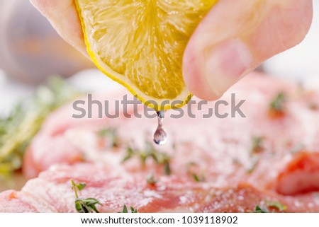 The man's hand squeezes the lemon juice with a raw pork steak. Also on the table, onion, thyme, garlic, salt, pepper, salt and pepper shaker on a wooden cutting board close-up Royalty-Free Stock Photo #1039118902