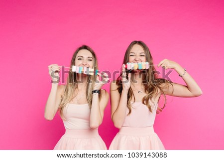 Studio portrait o two beautiful and sweet girls, in pink sunglasses and looks like princes or candies, sending kiss at camera. Pretty women showing marshmallows on stick and having fun. Fashion.
