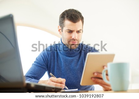 Young man wearing turtleneck sweater while using digital tablet and doing some paperwork at home. Home office.