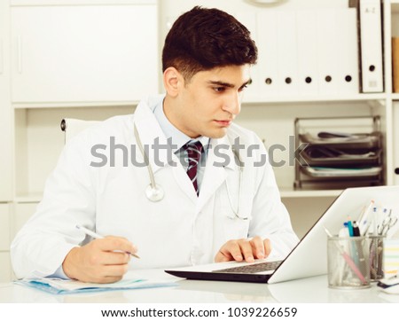 Male doctor 28-35 years old is doing report about patients in hospital. Royalty-Free Stock Photo #1039226659