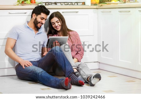 Joyful lovely couple browsing pictures on the tablet, sitting on the kitchen floor, having warm and nice talk