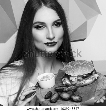 Portrait of beautiful young woman holding round tray with delicious burger, salad and fried potato lying on piece of kraft paper. Elegant female model demonstrating cheeseburger on wooden plate.