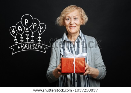 Wonderful present. Kind cheerful aged woman smiling and looking pleased while holding a beautiful red box with a birthday present for her beloved husband