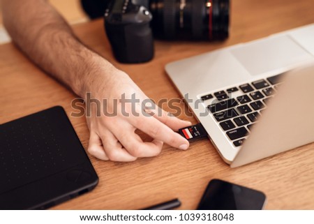 Close up freelancer bearded man in t-shirt using memory card at laptop sitting at desk.