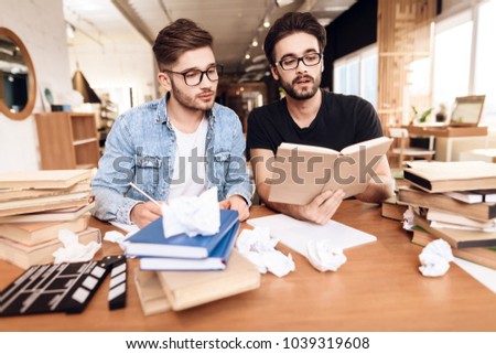 Two freelancer men in jeans shirt and t-shirt reading notes at laptop at desk surrounded by books.