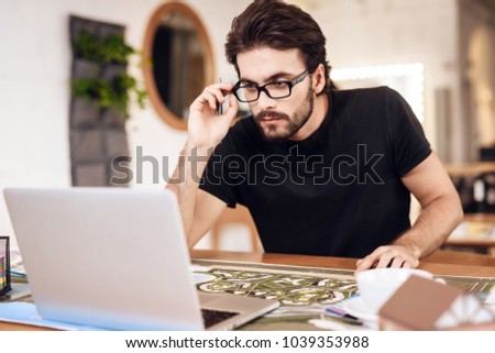 Freelancer bearded man in t-shirt looking at laptop sitting at desk.