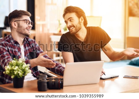 Two freelancer men in shirt and t-shirt discussing at laptop at desk.