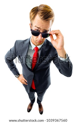 Are you seriously?! Full body portrait of funny skeptic young businessman in black confident suit and red tie, looking through sunglasses, top angle view shot, isolated against white background. 
