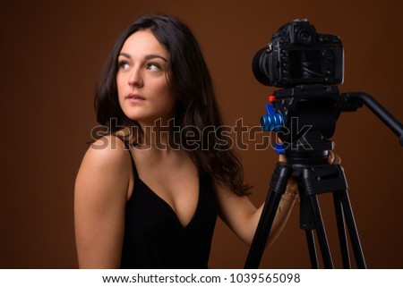 Studio shot of young beautiful woman vlogging while wearing black sleeveless top against brown background