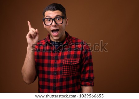 Studio shot of young handsome man wearing red checkered shirt with eyeglasses against brown background
