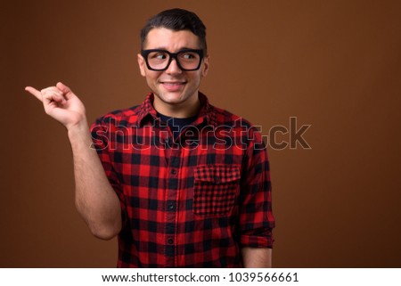 Studio shot of young handsome man wearing red checkered shirt with eyeglasses against brown background