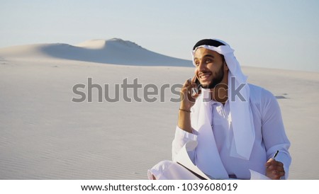 Handsome Arabian UAE Sheikh male businessman talks on mobile with business partner and solves important business issues and problems, holds paper project and makes notes with ballpoint pen, sitting on