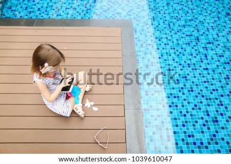 little kid girl taking selfie on tablet sitting close to blue swimming pool wearing flower and hold toy. concept of new technology or travel recreation, holiday family time on sunny day