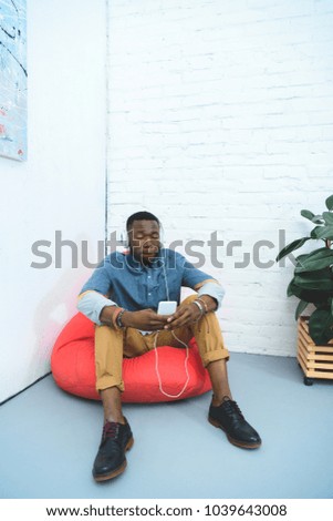 Young man with smartphone in hands listening to music in earphones while sitting in bean bag
