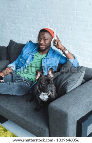 Frenchie dog on sofa sitting by African american man in headphones listening to music 