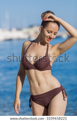 Portrait of attractive girl on beach. Beautiful young woman in brown bikini posing with arm raised at the sea in morning time