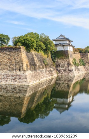 A classic white guard tower and the giant stone walls of Osaka castle are reflected in the calm waters of the wide outer moat in Osaka, Japan.