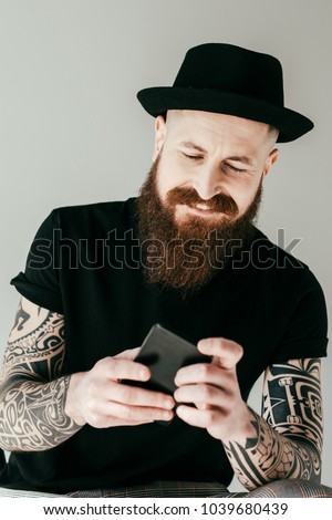 smiling handsome bearded tattooed man looking at smartphone isolated on grey