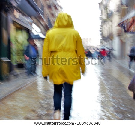 Impressionist photo at very low speed of blurred human figures in movement, on a rainy day, with umbrellas and raincoats, camera trepidation on purpose to give a sense of urgency, instability, hurry,