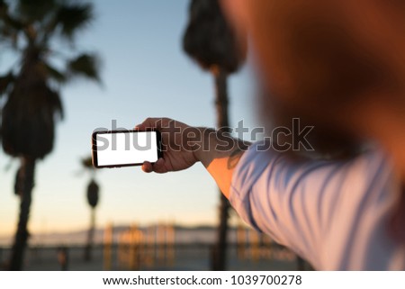 Cropped image of male's hand holding smartphone with mock up screen taking selfie outdoors, close up back view of man using modern telephone camera and app for making picture of evening landscape