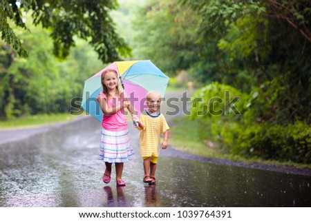 Kids playing out in the rain. Children with umbrella play outdoors in heavy rain. Little girl and boy caught in first spring shower. Kids outdoor fun by rainy autumn weather. Child in tropical storm.