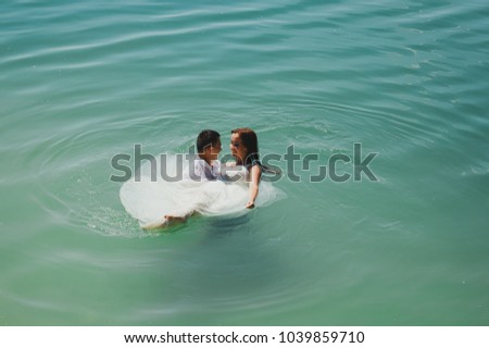 Wedding couple is hugging in azure blue lake splashes of water. Beautiful bride in puffy dress and groom are having fun. Summer passion crazy emotions photo on the seaside. Wet wedding clothes.