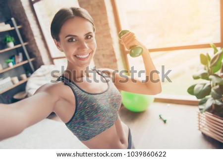 Close up portrait of delightful beautiful ideal slim sportive powerful muscular positive woman dressed in tight gray top demonstrating her biceps taking selfie having video call