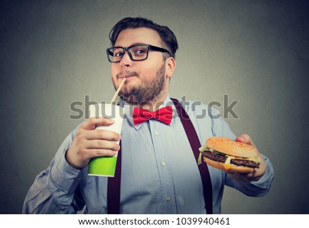 Young chunky man in formal outfit enjoying soda and hamburger on gray background. 