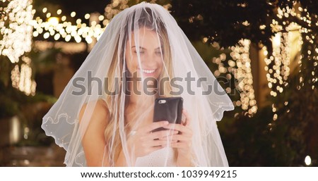 Young bride in wedding dress and veil taking a selfie with cell phone outdoors