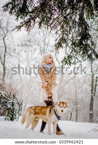 A beautiful blonde in a fur coat stands in the park with her dog