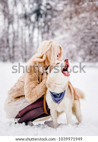 Blonde in a fur coat sitting with her dog in a winter forest