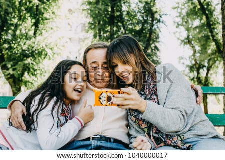 
A father with his daughters sitting on a stone bench, taking themselves some photographs. Father's day. Relaxed autumn day in family outdoors. Lifestyle portrait.

