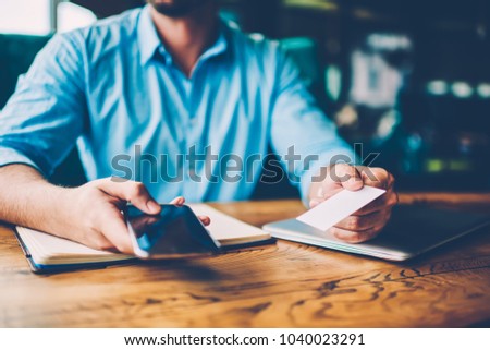 Cropped image of hands of stylish businessman sitting at wooden desktop in coffee shop interior with laptop computer and notepad holding modern digital telephone device and business card in hands