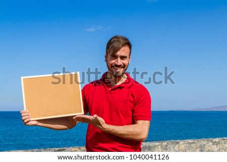 Bearded attractive inviting young man holding a framed blank corkboard in blue sky